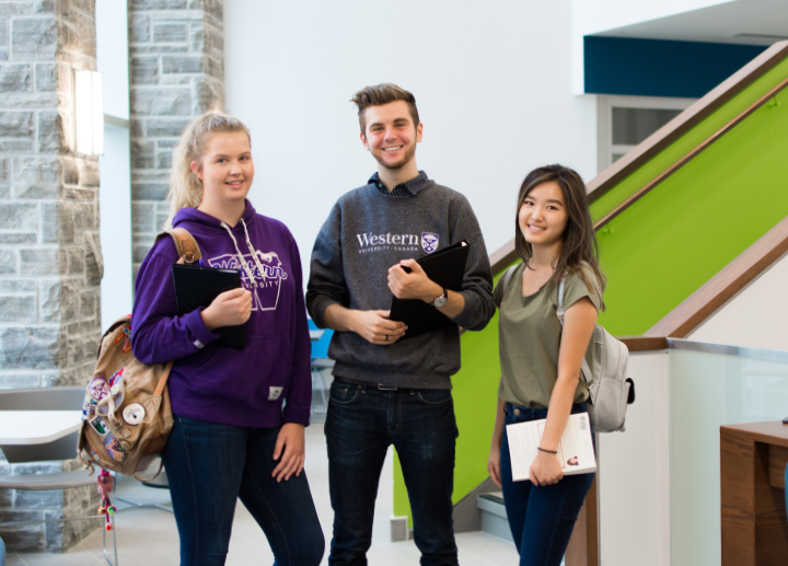 Three university students smile indoors with books and backpacks near a green stair railing.
