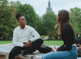 Two students chatting on a picnic table on a fall day on campus.