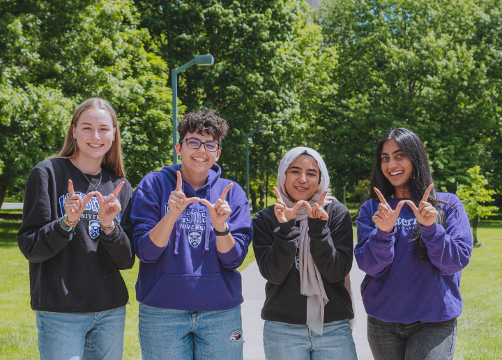 Four students, smiling for a photo on campus, holding up W's with their hands for Western.